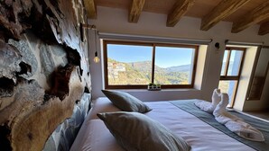 Une chambre dotée d’une grande fenêtre donnant sur un paysage montagneux. La pièce est agrémentée de poutres apparentes et d’un mur en pierre rustique.