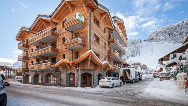 Un immeuble en bois à plusieurs étages, doté de balcons, avec un télésiège en arrière-plan et un paysage enneigé.