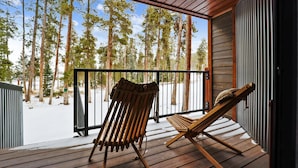 A wooden deck with two Adirondack chairs, a metal railing, and a view of tall pine trees.