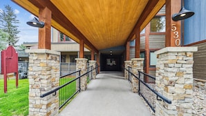 A covered walkway with stone pillars and a wooden ceiling, leading to a building entrance.