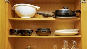 A wooden cabinet with shelves containing kitchenware, including a colander, bowls, cups, plates, a glass jar, and wine glasses.