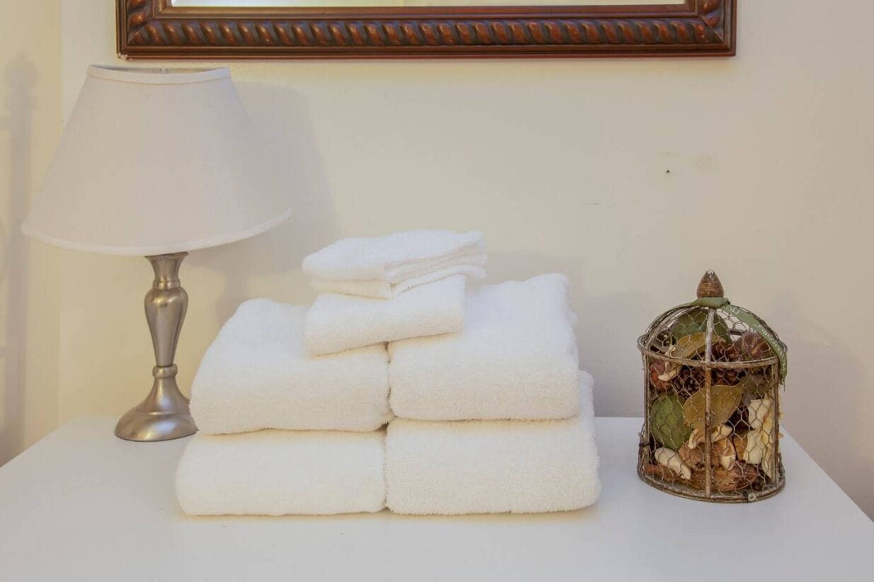 Een stapel witte handdoeken, een lamp en een decoratieve vogelkooi op een witte tafel.