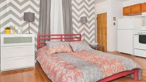 Une chambre à coucher comprenant un lit, une table de chevet, une lampe, une commode et un coin cuisine avec un réfrigérateur.