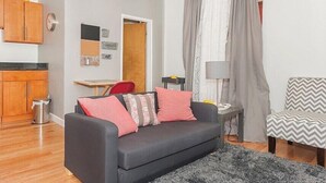 Un espace de vie compact comprenant un canapé, un fauteuil, une petite table et une lampe. On y trouve également un meuble en bois et une poubelle.
