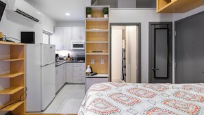 Une chambre d’hôtel de taille réduite, comprenant un lit, une kitchenette, une petite salle de bain et une porte donnant sur une autre pièce.