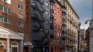 Un immeuble à plusieurs étages doté d’une échelle de secours, d’une enseigne sur laquelle on peut lire « ORGIAN IN THE HILL », et de voitures garées dans la rue.