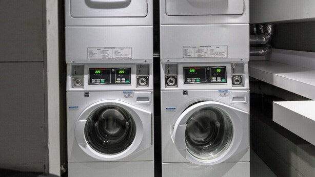 Deux machines à laver à chargement frontal, superposées, se trouvent dans une buanderie, avec des panneaux de commande visibles et des tuyaux apparents au-dessus.