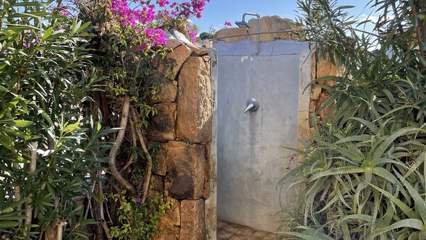 A stone-walled shower area with a metal door, surrounded by lush greenery and flowering plants. - Holiday Home just 1 km far from Porto Ainu Beach  (Sardegna)