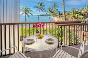 A balcony with a table set for two, overlooking a beach with palm trees and the ocean.