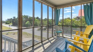 En overdækket veranda med udsigt til parkeringsplads og grønt, med gule og blå Adirondack-stole og en lille hvid stol.