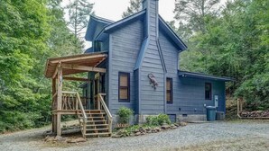 A blue wooden house with a steep roof, a wooden porch, and a stone chimney. - Easy Access Cabin deep in the trails but very close to the city! (Murphy)