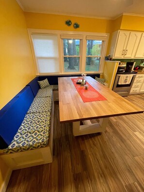 A compact kitchen with a wooden table, a built-in bench with patterned cushions, and a window with a view of trees.