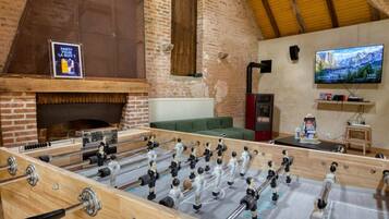Une table de baby-foot dans une pièce rustique avec une cheminée en briques, un téléviseur et un confortable canapé vert.