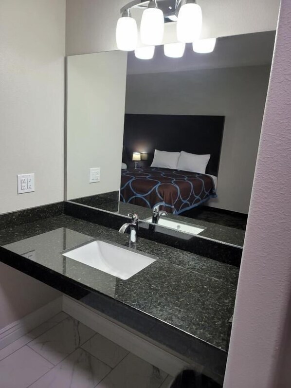A modern bathroom with a large mirror, a granite countertop, a white sink, and a bed with a patterned bedspread.