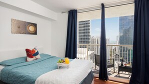 Ein modernes Hotelzimmer mit Balkon, einem Bett mit blauer Bettdecke, einem kleinen Tisch mit Obst und Saft sowie Blick auf die Stadt.
