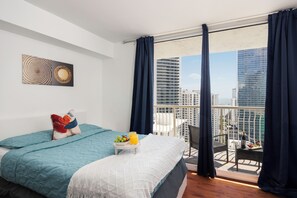 A modern hotel room with a balcony, a bed with a blue bedspread, a small table with fruit and juice, and a city view.