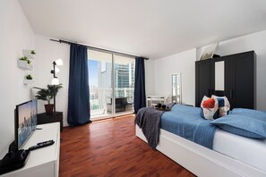 A modern bedroom with a balcony, a bed with blue bedding, a desk with a TV, and a potted plant.