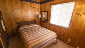 A bedroom with a wooden floor, a bed with a striped blanket, a nightstand with a lamp, a window with blinds, and a wooden ceiling. - Condo in the mountains! (Red River)