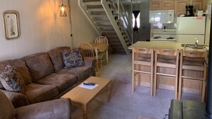 Ein gemütliches Wohnzimmer mit einer braunen Ledersofa, einem Holztisch und einem Essbereich mit Holzstühlen. Eine Treppe führt zu einer Küche mit Mikrowelle und Herd.