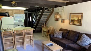Ett mysigt vardagsrum med en brun lädersoffa, trästolar, ett soffbord och en köksdel med en spis och ett kylskåp.