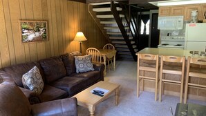 Een gezellige woonkamer met een bruine leren bank, een houten salontafel en een eethoek met een houten tafel en stoelen. Een trap leidt naar een verdieping erboven.