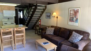 Een gezellige woonkamer met een bruine leren bank, houten stoelen, een klein houten tafeltje, een trap, een magnetron en een lijst met een foto aan de muur.