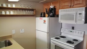 Een kleine keuken met een koelkast, magnetron, fornuis en een plank met bekers en glazen.
