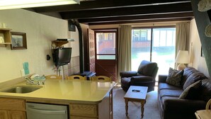 Cocina compacta con lavabo, estufa y televisor. Hay un comedor con mesa y sillas, y una sala de estar con sofá y lámpara.