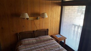 Un dormitorio con cabecera de madera, colcha a cuadros, dos lámparas de pared y una ventana con persianas.