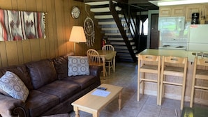 A cozy living room with a brown leather couch, patterned pillows, a wooden coffee table, and a dining area with wooden chairs and a table. A staircase leads to a kitchen with a microwave, coffee maker, and stove. - Condo in the mountains! (Red River)