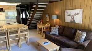 A cozy living room with a brown leather sofa, a wooden coffee table, and a dining area with a wooden table and chairs. A staircase with a wooden railing is visible in the background. - Condo in the mountains! (Red River)