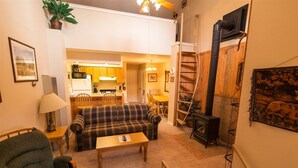 A cozy living room with a sofa, a coffee table, and a fireplace. There is a kitchen in the background with wooden cabinets and a dining area. - Condo in the mountains! (Red River)