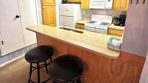 Une cuisine compacte avec des meubles en bois, un réfrigérateur blanc, une cuisinière et deux tabourets de bar noirs.