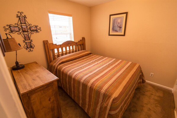 Une chambre à coucher avec un lit en bois, une table de chevet, une lampe, une fenêtre avec des stores et un tableau encadré au mur.