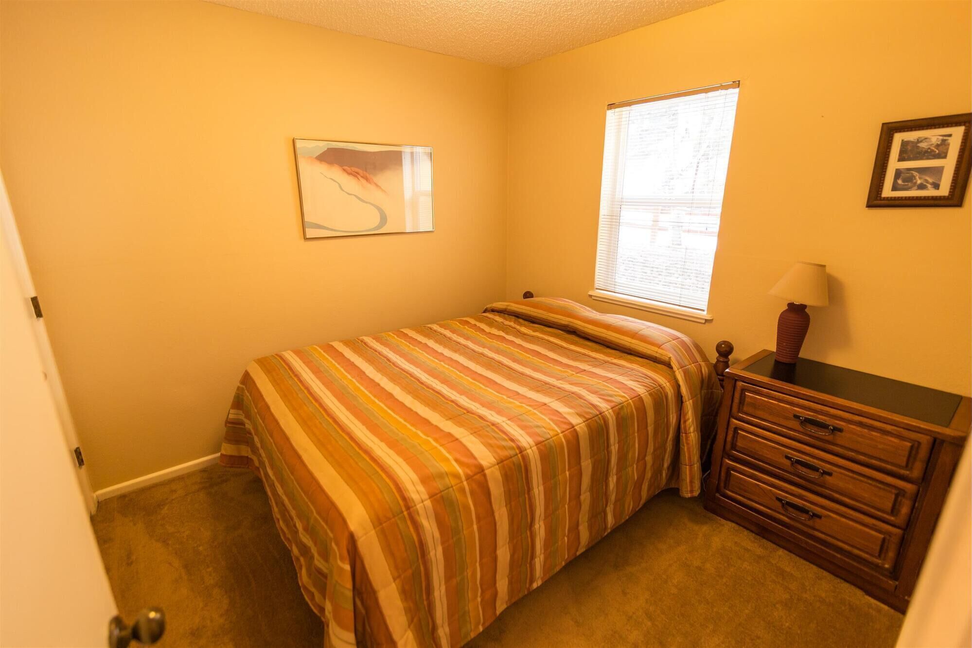 Une chambre à coucher comprenant un lit, une table de chevet, une commode, une fenêtre avec des stores et un tableau accroché au mur.