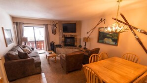 A cozy living room with a fireplace, a dining area with wooden chairs, and a large window with curtains. - Condo in the mountains! (Red River)