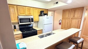 A compact kitchen with wooden cabinets, a microwave, stove, refrigerator, and a bar area with stools. - Condo in the mountains! (Red River)