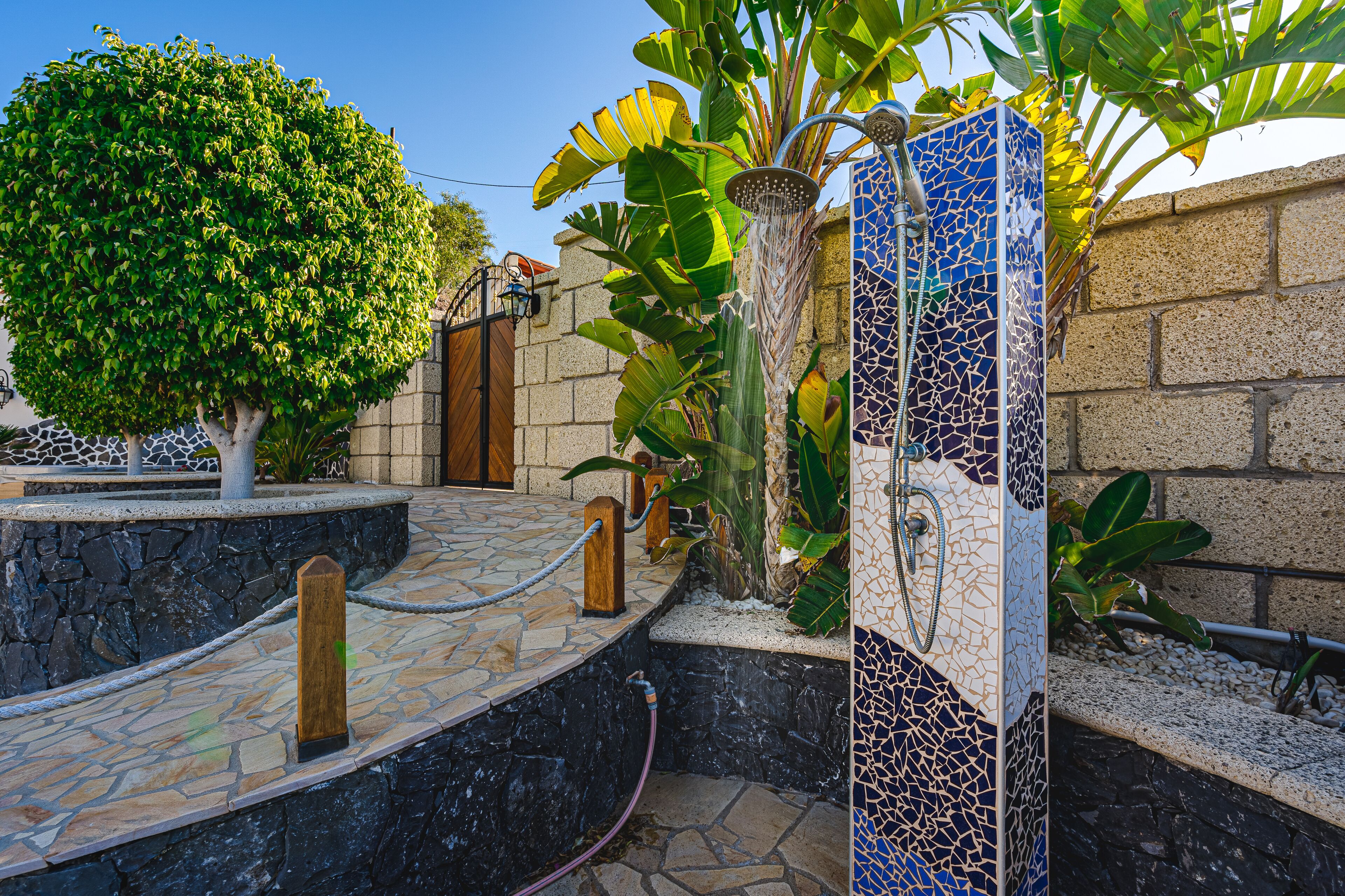 Un chemin de pierre mène à une zone de douche carrelée, dont le mur est décoré d’une mosaïque, le tout étant entouré de verdure et d’un mur en pierre.