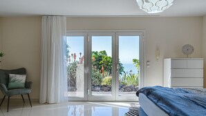 A bedroom with a bed, a chair, a white cabinet, and a view of a garden through a sliding door. - Luxury villa with fantastic sea views (Adeje)