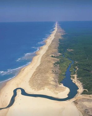 An aerial view of a coastal area with a long sandy beach, a river running parallel to the coast, and dense green vegetation on the land.