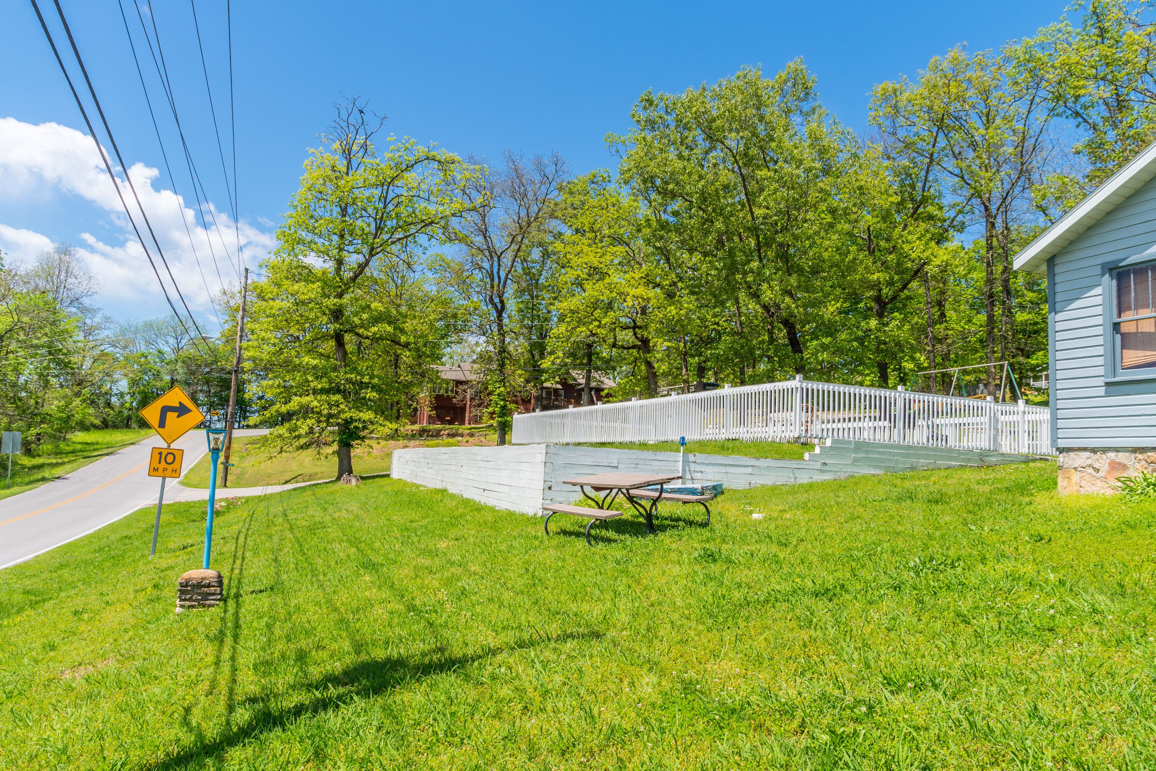 A residential area with a paved driveway, a picnic table, a white picket fence, and a house with a blue exterior.