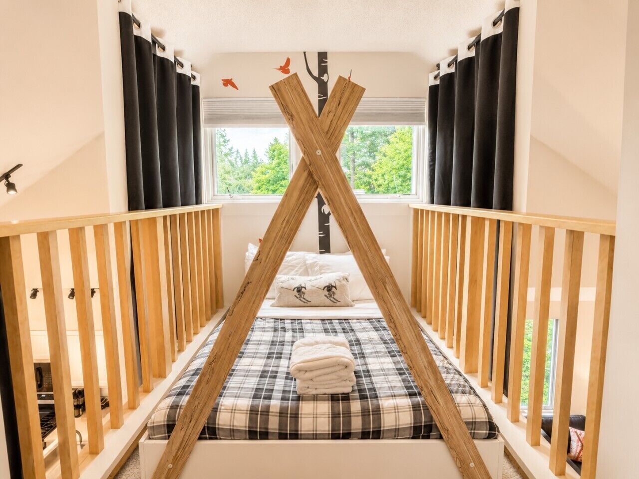 Ein Zimmer mit einem Bett, das unter einer hölzernen, kreuzförmigen Deckenkonstruktion zentriert ist, umrahmt von schwarzen Vorhängen und einem Fenster mit Blick auf Bäume.
