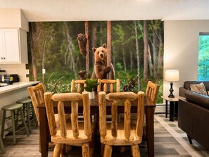 Un coin repas avec une table et des chaises en bois, une cuisine à l’arrière-plan et une fresque représentant une forêt sur le mur.