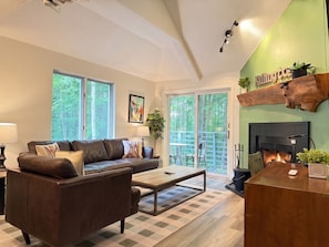 A living room with a sofa, coffee table, and fireplace. There is a large window and a balcony with outdoor furniture.