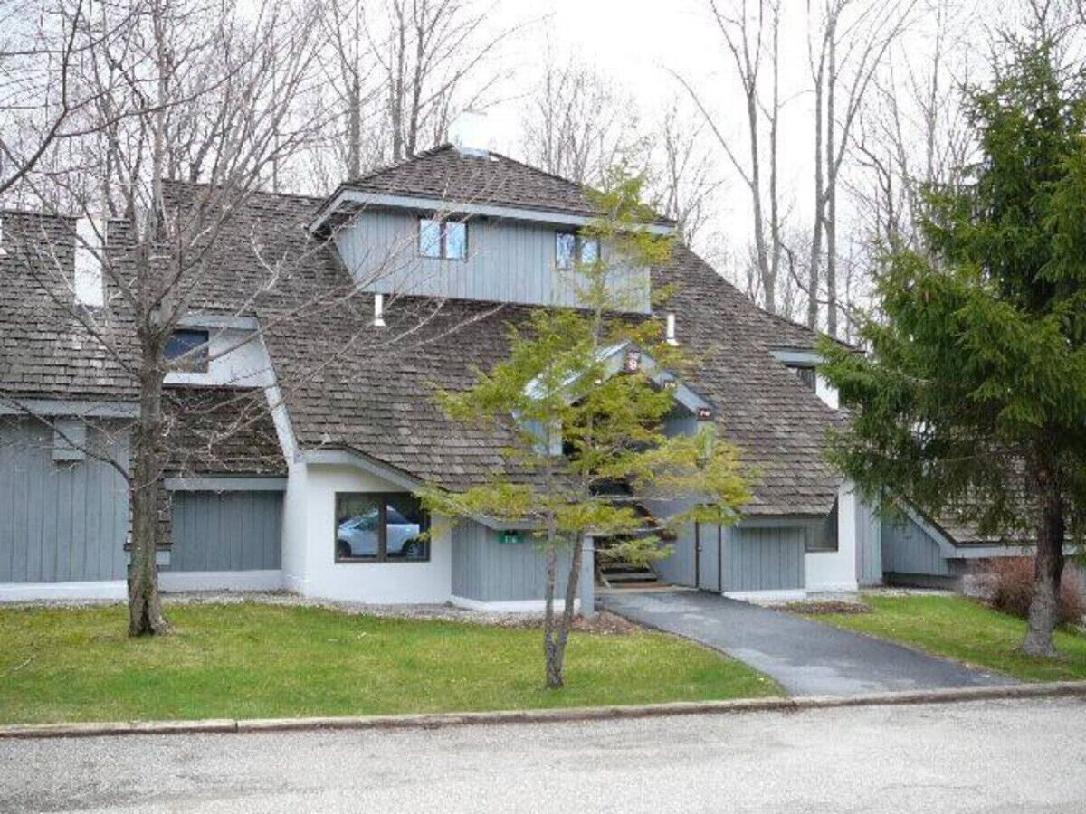 A two-story residential building with a sloped roof, a covered driveway, and a small front yard with a young tree.