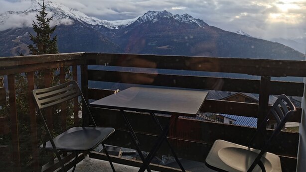A balcony with a table and two chairs, overlooking a mountainous landscape with snow-capped peaks and a clear sky.