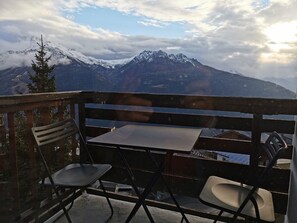 A balcony with a table and two chairs, overlooking a mountainous landscape with snow-capped peaks and a clear sky.