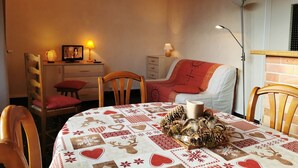 A cozy room with a round table covered in a festive tablecloth, a bed with a red blanket, and a desk with a lamp and picture frame.