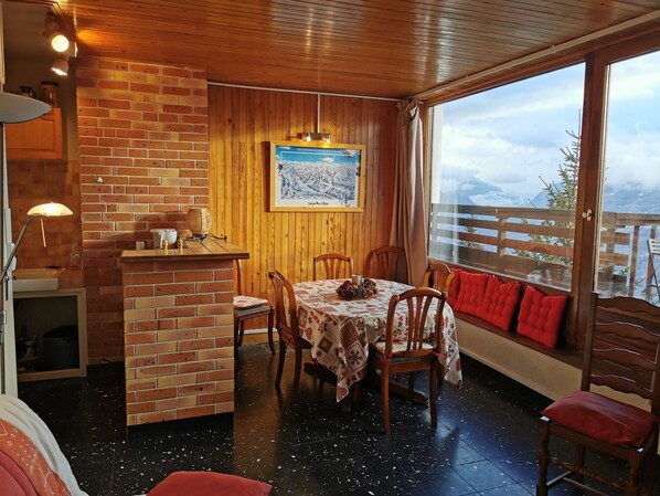 A cozy dining area with a brick wall, wooden ceiling, and a table set for four. A framed picture hangs on the wall, and a window offers a view of the outdoors.