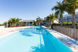 A swimming pool with a clear blue water, surrounded by a fence, palm trees, and a building in the background. - Stylish apartment in the North with Teide views (Santa Úrsula)